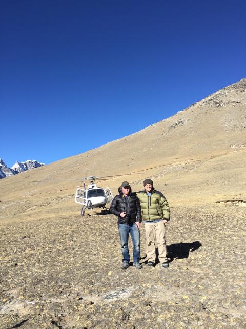 Two men in down jackets pose on rocky high-altitude ground in Nepal, a helicopter parked behind them below snowy peaks