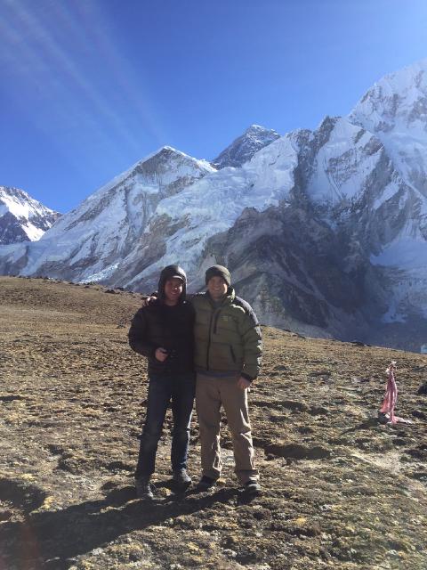 Two smiling men in hats and puffy jackets stand arm in arm before massive snow-covered Himalayan peaks near Everest