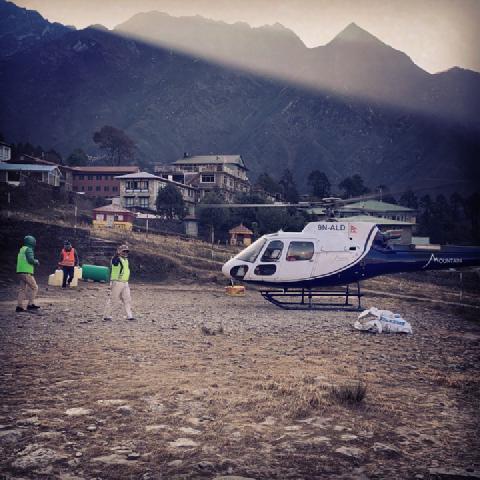Workers in safety vests carry supplies past a helicopter on a gravel helipad in a Nepali mountain village at dusk