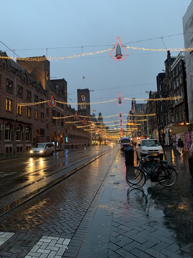 Rainy Amsterdam street at dusk with strings of holiday lights overhead, wet tram tracks, and a parked bicycle on the brick sidewalk