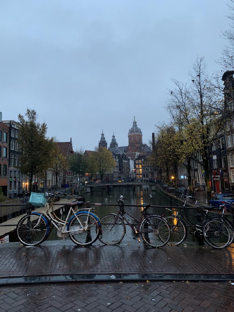 Bicycles lean on a bridge railing over an Amsterdam canal, with the domed Basilica of St. Nicholas rising in the grey distance