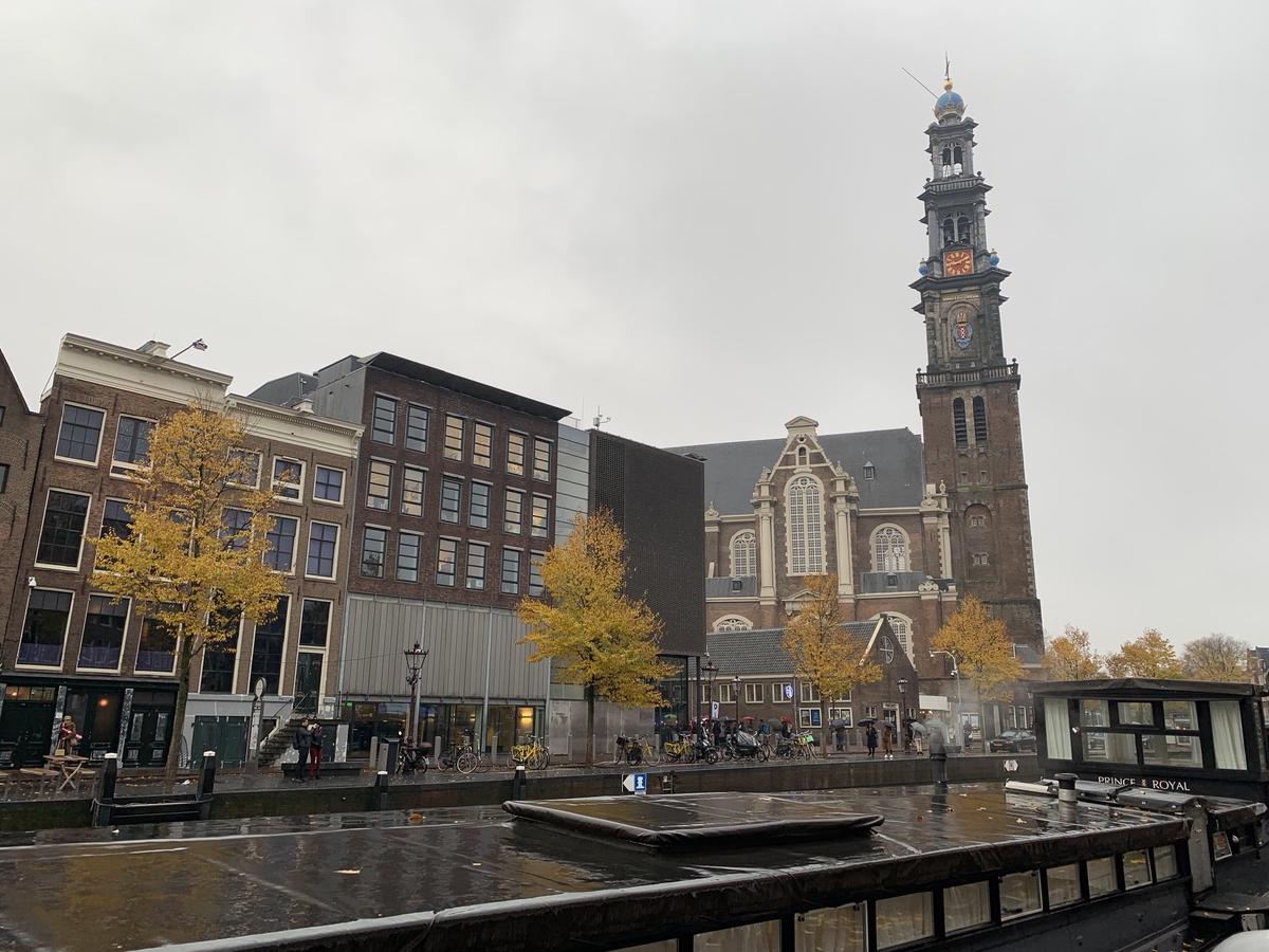 The tall clock tower of Amsterdam&rsquo;s Westerkerk above canal houses and autumn trees, seen across the water on an overcast day