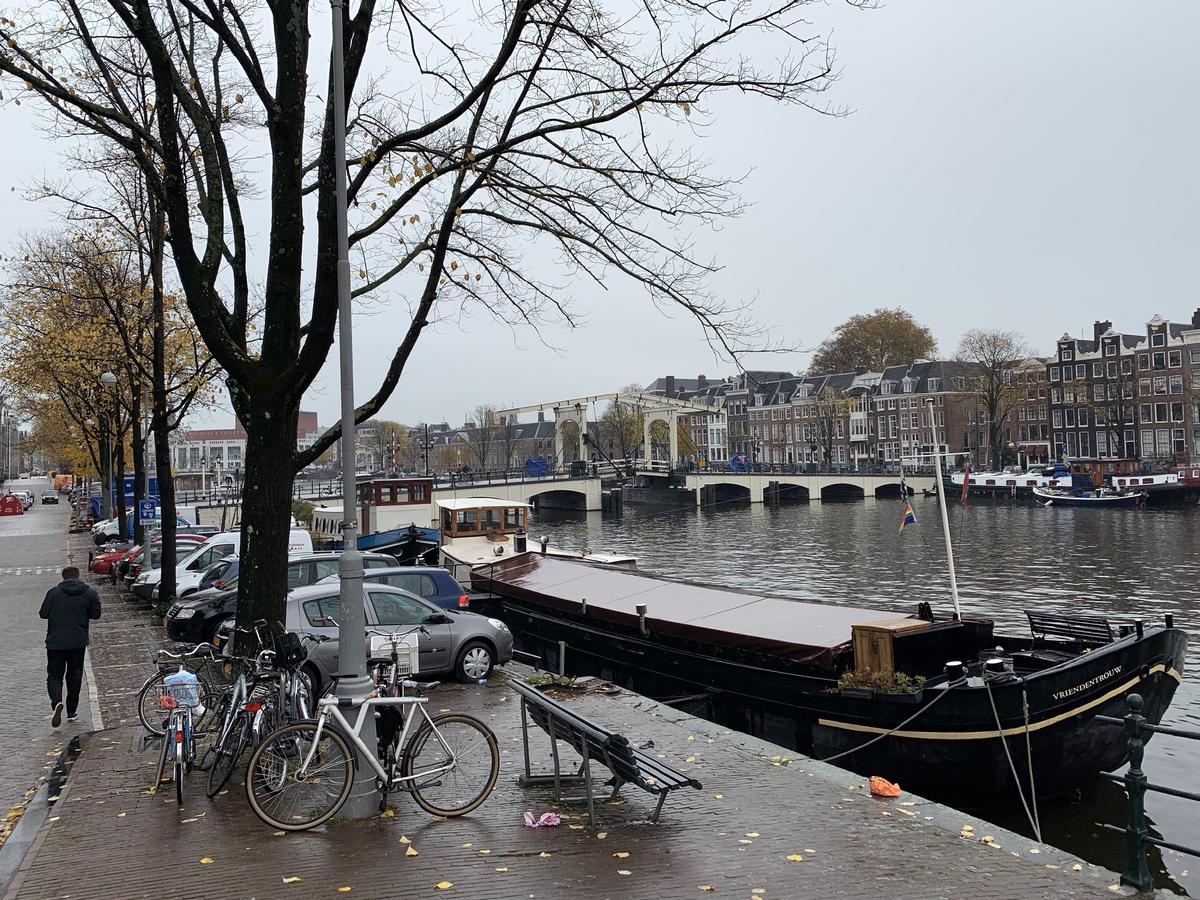 Black houseboat moored on the Amstel in Amsterdam near a white drawbridge, with bikes and bare autumn trees along the quay