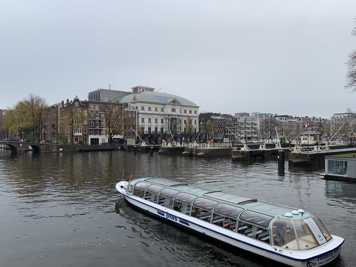 Glass-roofed canal tour boat motors along the Amstel in Amsterdam past river locks and the domed Royal Theater Carre