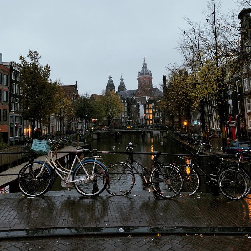 Weathered bicycles chained along a wet Amsterdam bridge at dusk, canal and the towers of St. Nicholas Basilica beyond