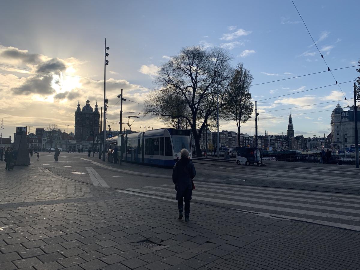 Person crossing a broad brick plaza in Amsterdam as a blue-and-white tram passes, church towers silhouetted against the low sun