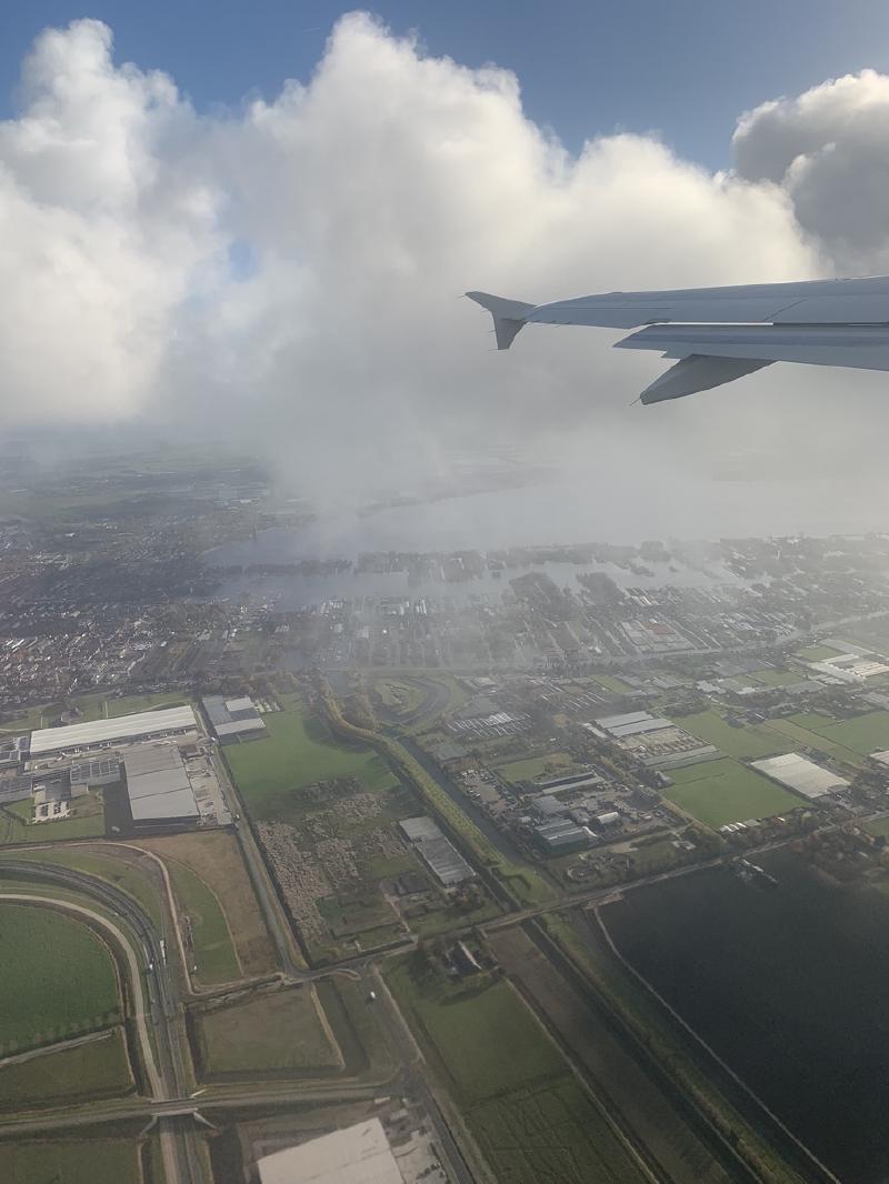 Airplane wing over Dutch farmland, greenhouses, and waterways near Amsterdam, low clouds drifting below