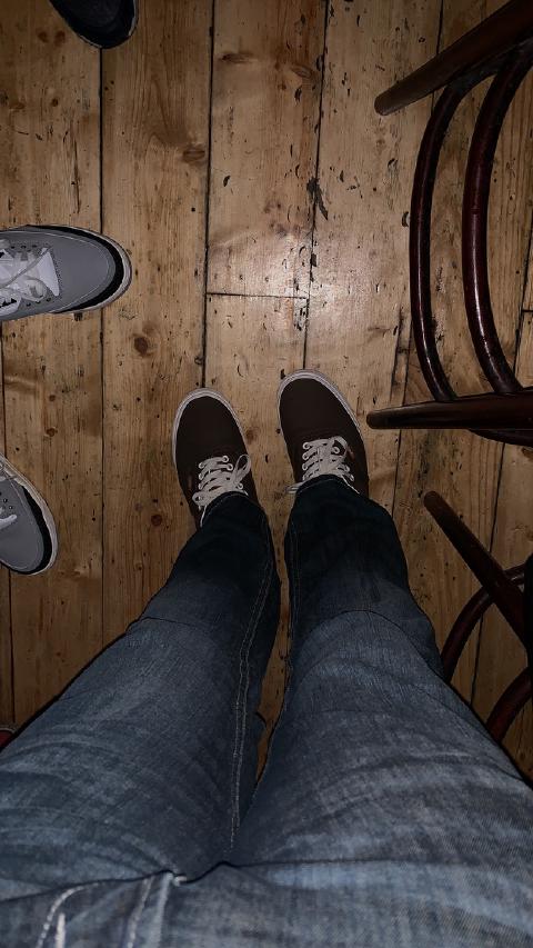 Looking down at brown sneakers and jeans on a worn wooden bar floor, another pair of grey shoes and chair legs at the edges