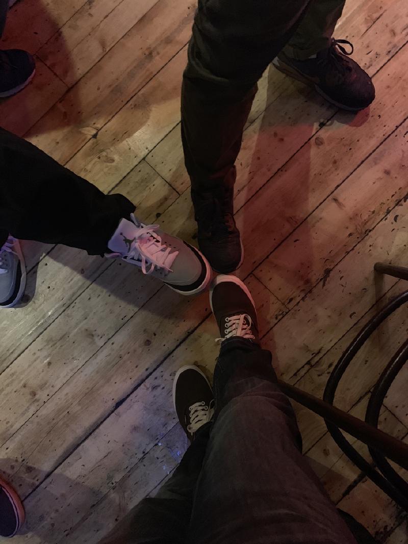 Several friends’ sneakered feet gathered in a loose circle on a scuffed wooden floor under dim red bar light