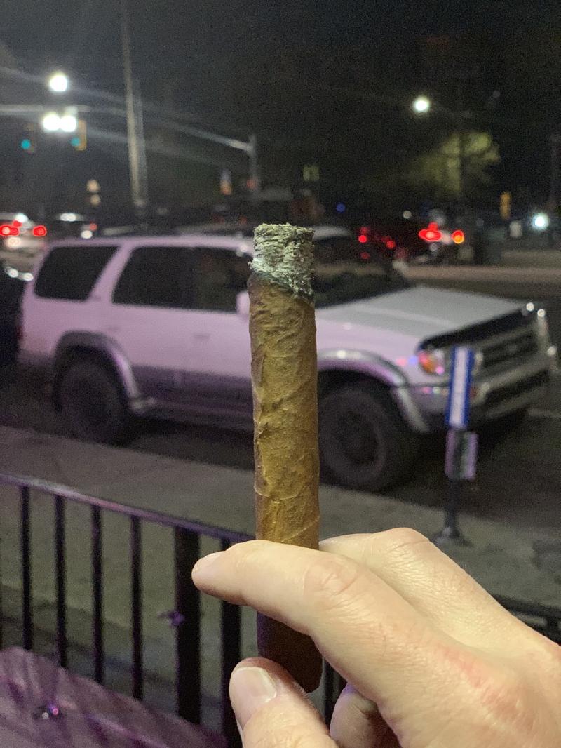 Hand holds up a half-smoked cigar with long grey ash at night, a white SUV and street lights blurred behind
