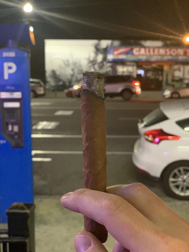 Cigar with ashed tip held up at night beside a blue parking kiosk, storefront sign and parked cars out of focus across the street