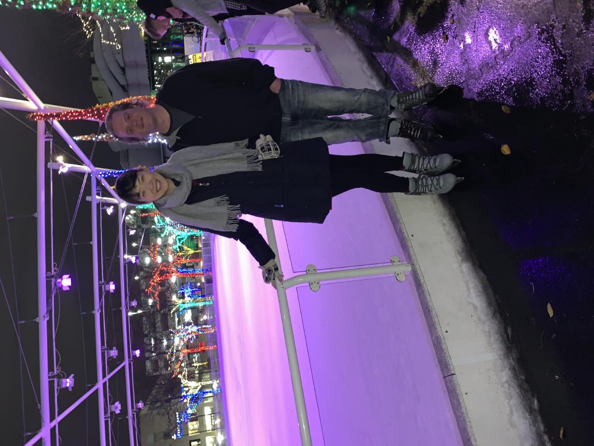 Man and woman in skates pose at the rail of an outdoor ice rink at night, trees wrapped in colorful holiday lights behind them