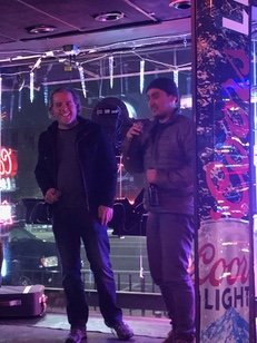 Two men share a laugh at an outdoor night event, one holding a microphone beside a Coors Light-wrapped column