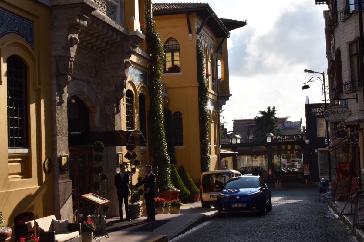 Doormen in suits chat outside an ivy-draped yellow Ottoman-style hotel on a cobbled Istanbul street, cafe signs down the block