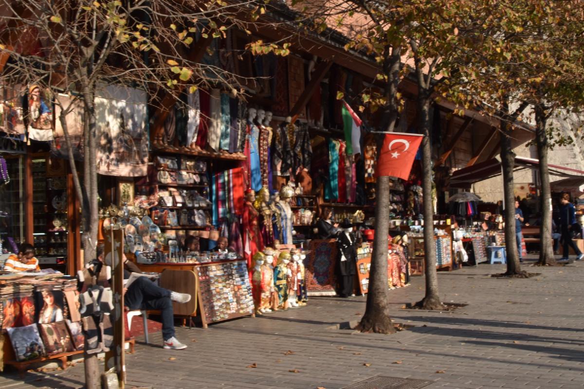 Bazaar stalls in Istanbul selling scarves, kaftans, magnets, and dolls under autumn trees, a Turkish flag hanging out front