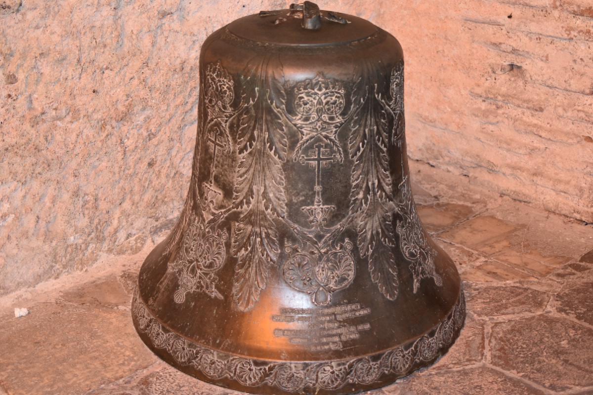 Large bronze bell engraved with crosses and leaf garlands sits on a stone floor against a rough brick wall