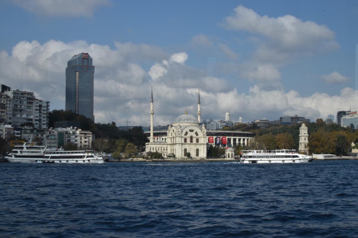 Ornate white Dolmabahce Mosque and its twin minarets on the Bosphorus shore, ferries passing and a high-rise tower behind