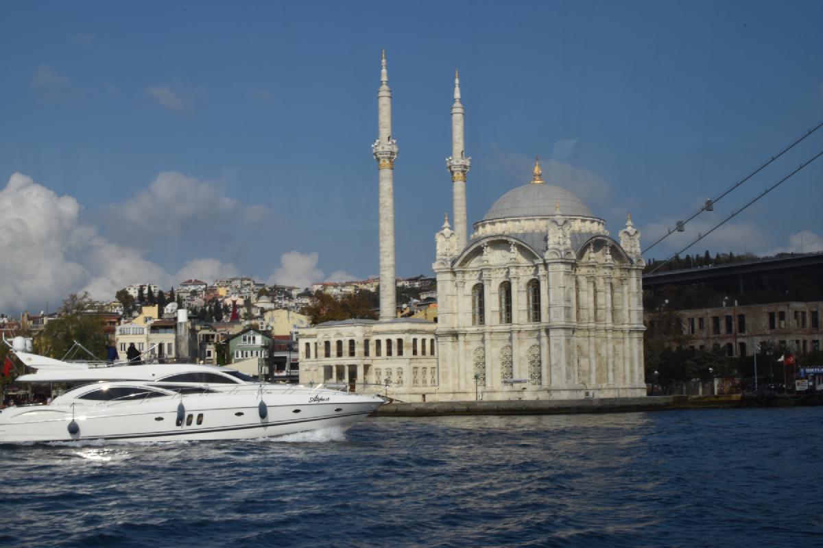 White motor yacht speeds past the ornate Ortakoy Mosque on the Bosphorus, the Bosphorus Bridge deck overhead at right