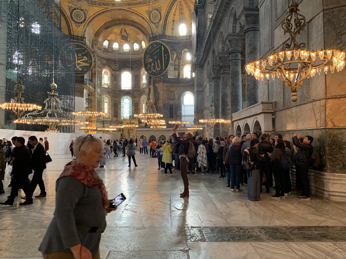 Visitors wander the marble floor of Hagia Sophia beneath glowing chandeliers, calligraphy medallions, and stained glass