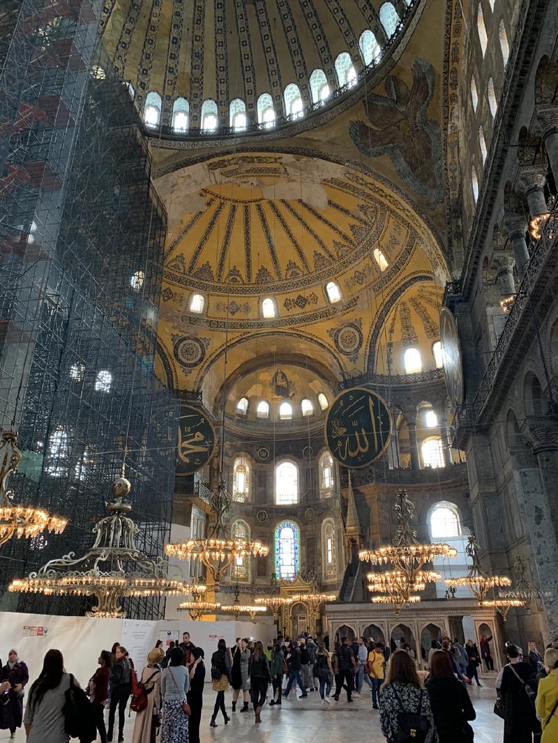 Crowds beneath Hagia Sophia&rsquo;s stacked golden domes, with a Madonna and Child mosaic in the apse and tall scaffolding at left