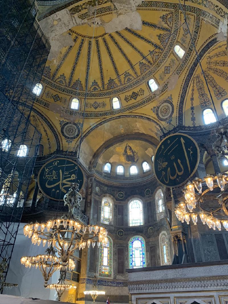 Hagia Sophia&rsquo;s painted domes and stained-glass windows above the marble mihrab, brass chandeliers hanging in the foreground