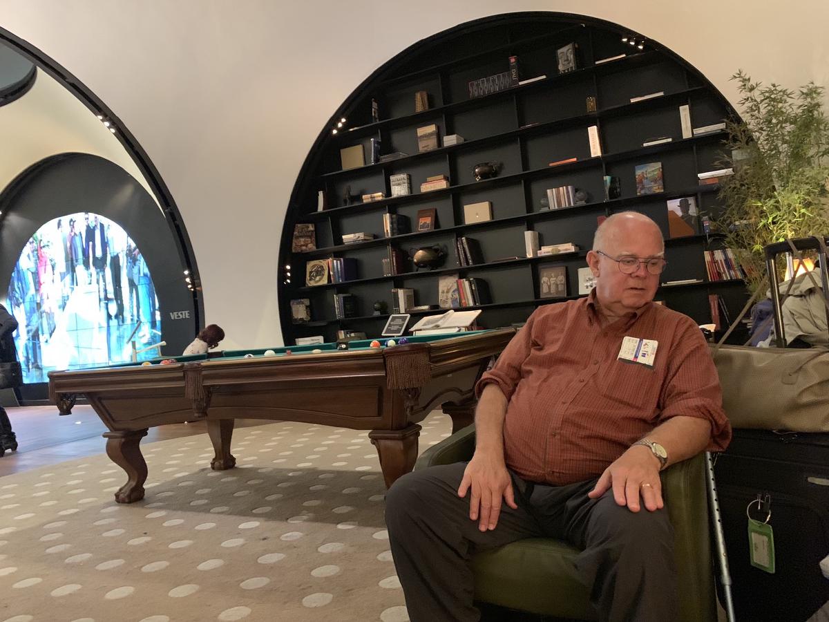 Older man rests in a green leather chair beside luggage in an airport lounge, pool table and round bookshelf wall behind him